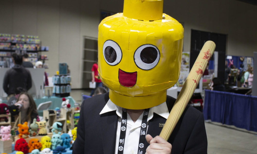 Photo Contains: A man holding a tiny baseball bat dressed in a suit while wearing a large yellow lego-like helmet.