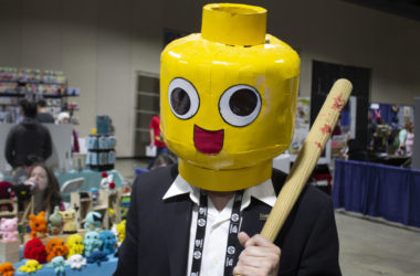 Photo Contains: A man holding a tiny baseball bat dressed in a suit while wearing a large yellow lego-like helmet.