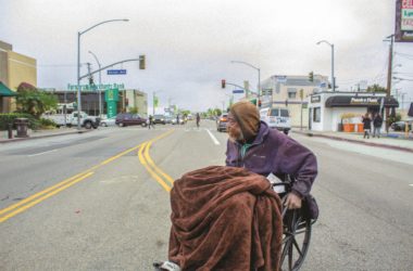 A man in a wheelchair crosses the street by passing through the middle of the road.