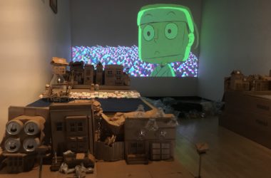 A cardboard crafted house sits in front of a screen where an animation takes place.