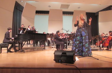 Chloë Agnew stands on a stage in a blue and b;ack dress in from of many students.