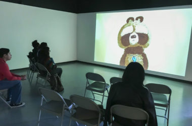 Students sit in a dark room to watch an animated film with a cartoon tanuki on the screen.