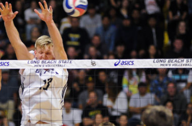 The upper part of Simon Andersen's body as he blocks a volleyball coming over the net. His face is scrunched and his arms are stretched far above his head.