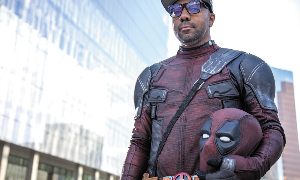 A man wears a deadpool costume while holding his mask in left arm. He's wearing a gray baseball cap and glasses. He is pictured from his waist up.