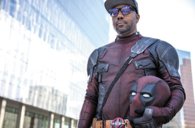 A man wears a deadpool costume while holding his mask in left arm. He's wearing a gray baseball cap and glasses. He is pictured from his waist up.