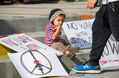 A young child sits on a curb surrounded by protest signs. One sign has a peace symbol and another says no war with Iran.