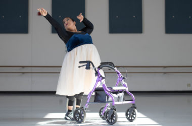 A young woman stands with her arms outstretched in a dance position next to a purple mobility aid. She is wearing a long tulle skirt.