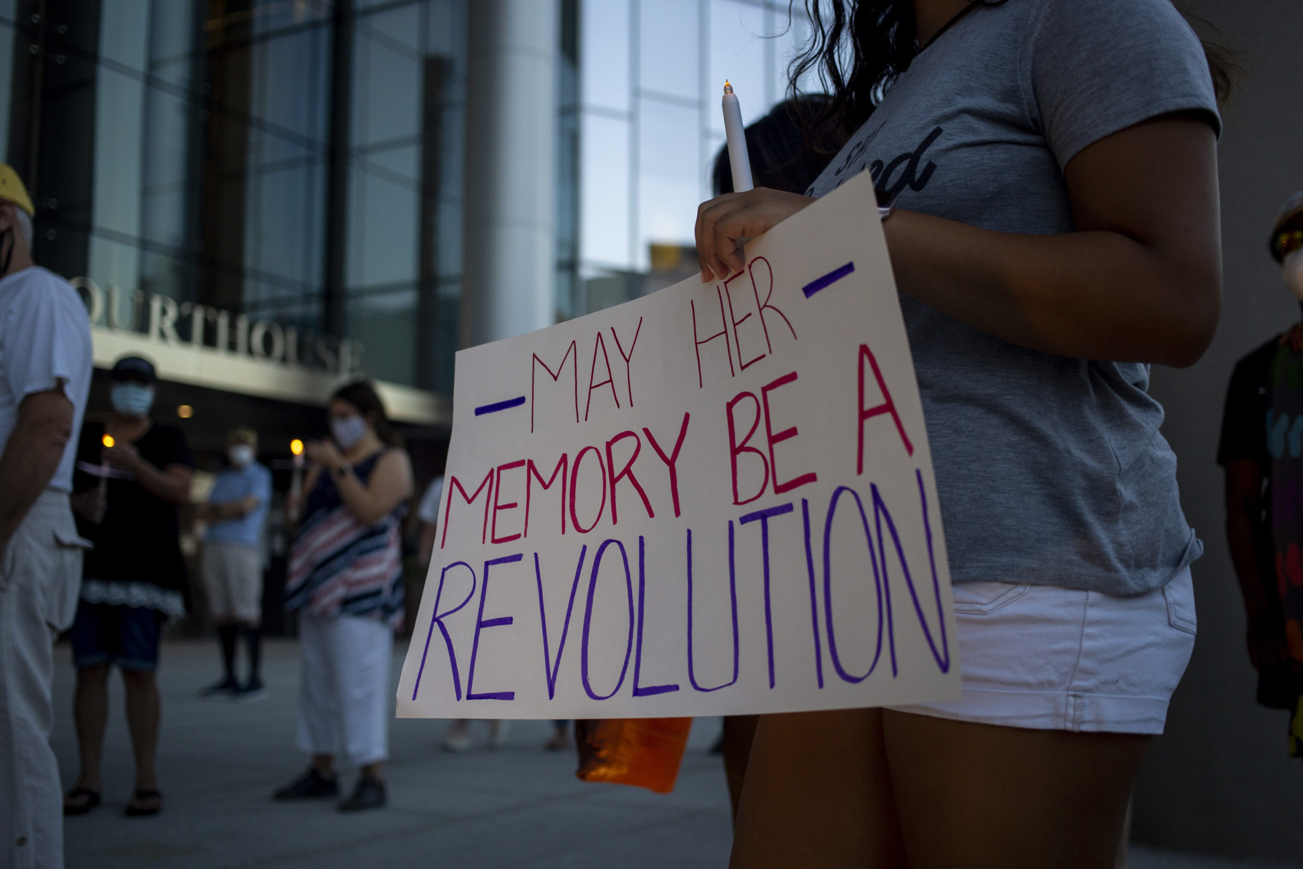 A woman holds a sign that reads "May her memory be a revolution"