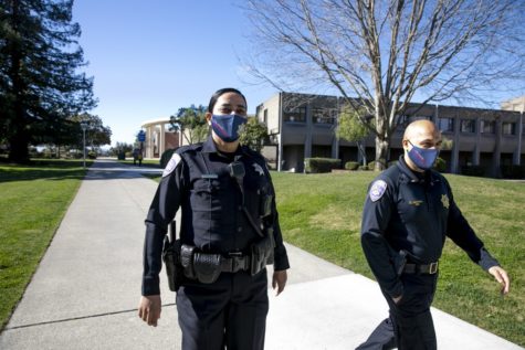 Two police officers with face masks walk on a college campus. One is male and one is female.