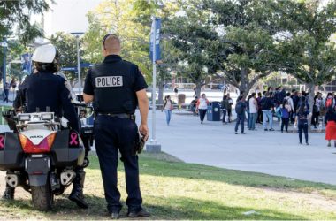 Two police officers watch a crowd on a college campus one sits on a motorcycle