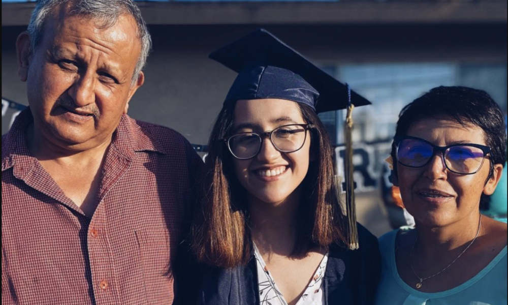 A father and mother stand with their daughter in the middle of them. The daughter is wearing a graduation gown and cap.