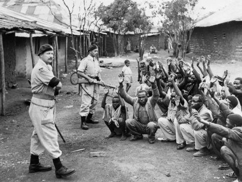 British authorities guard a group of men while searching for evidence that they participated in the Mau Mau Uprising.