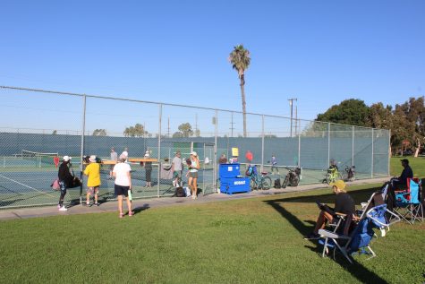 Intrigued spectators watch and people wait their turn to enter the pickleball courts to play the game.