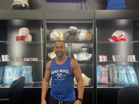 Raúl E. Anaya III, owner of Hard Worx Fitness standing in front of the trophies case on the right-hand side of the entrance. These trophies (belts) behind Anaya were won by the members in their competitions in the tournaments that the gym held.