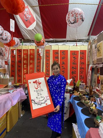 Khánh Diệp, a vendor and calligrapher at the Asian Garden Mall Tết Flower Festival posing with her art. Her scroll reads: Wealth and money, New year brings happiness with friends and family, Happy life, Health, Prosperity.
